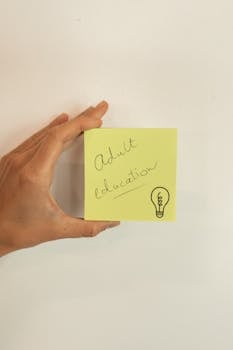 A hand holding a yellow sticky note featuring the words 'Adult Education' with a lightbulb icon.