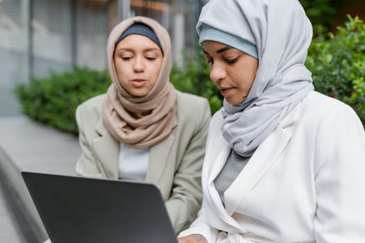 Two women in hijabs collaborate on a laptop outdoors, symbolizing friendship and teamwork.